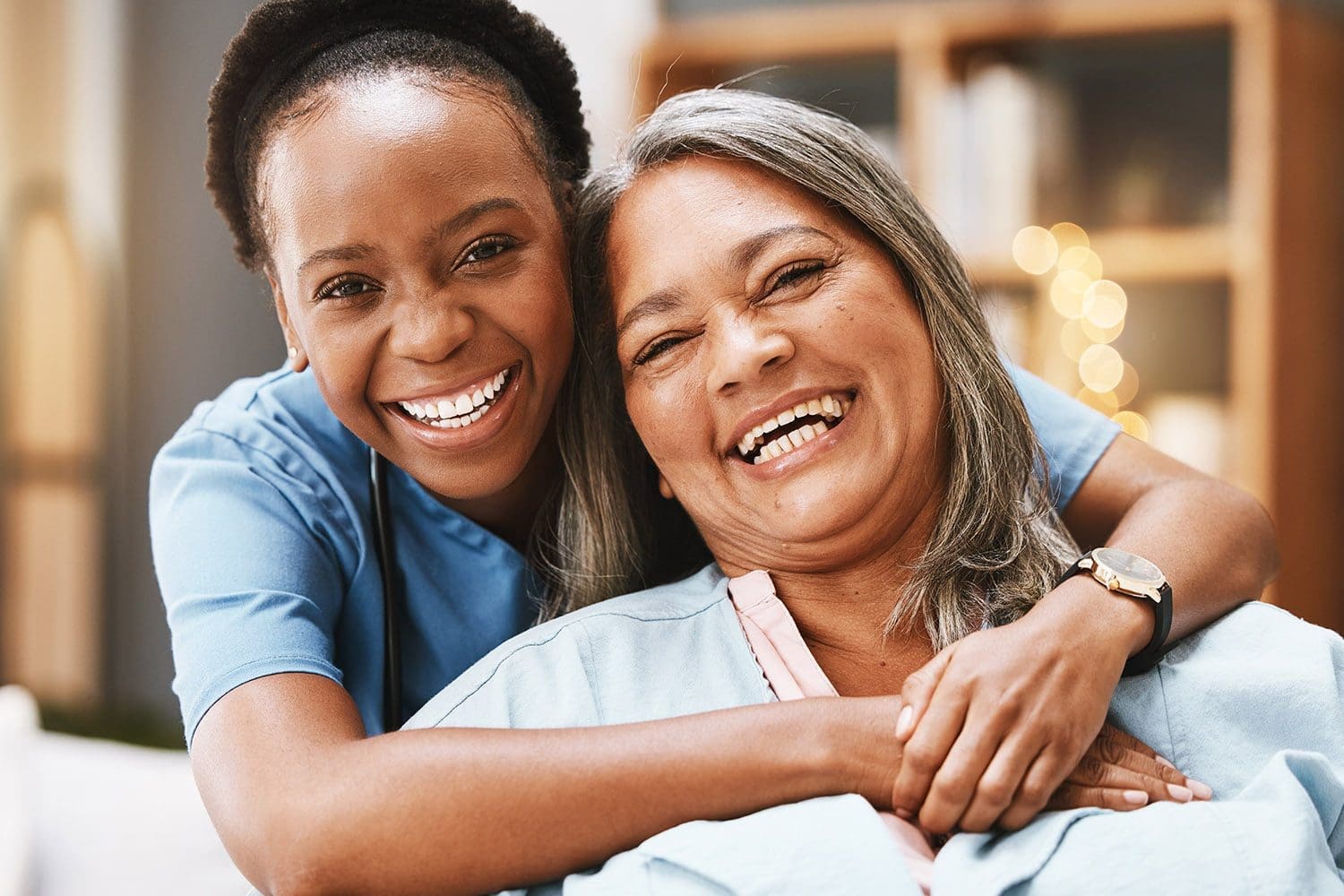 A black female carer hugs a dark skinned elderly female while both are smiling