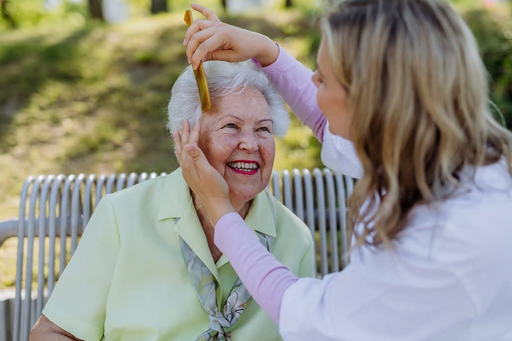 A carer helping senior woman to comb hair and make hairstyle when sitting on bench in park in summer.