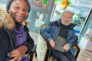 On the left is carer, Tamu, a black woman with dark hair wearing a grey winter hat, a purple Heritage Healthcare shirt and a black fluffy coat. To the right of her is client, Philip and old man with short grey hair and a grey beard. He is wearing a blue and black coat with a black hoodie underneath.