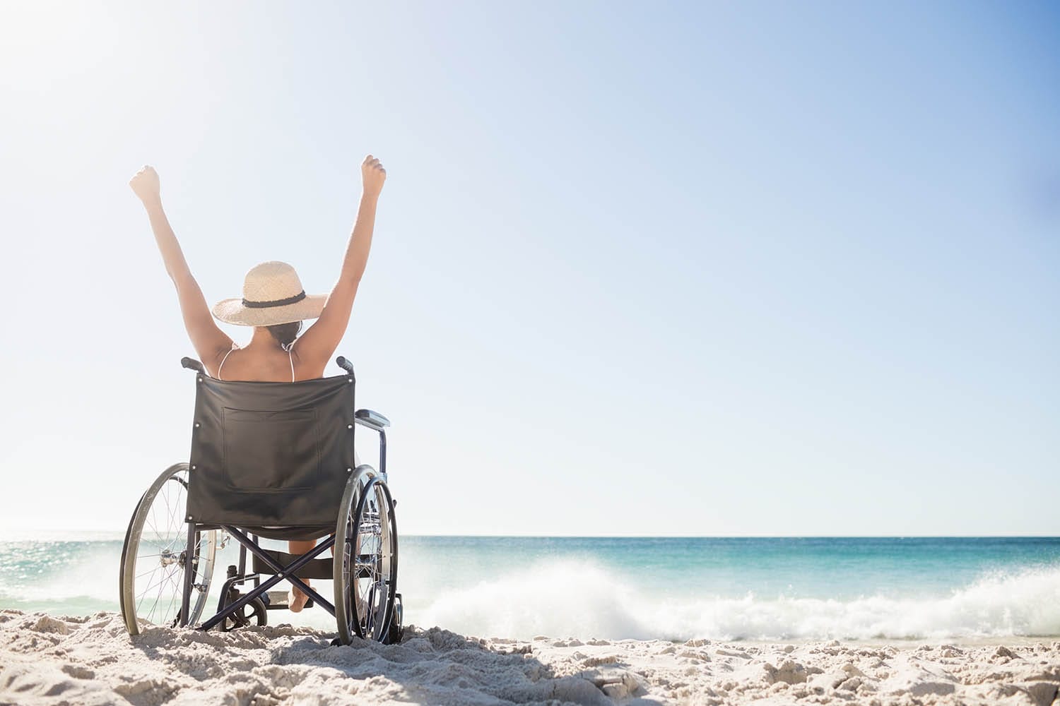 A white woman in a wheelchair, wearing a summer hat, with her arms raised on the beach