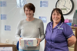 Carer, Caroline, a white women with short brown hair, wearing a grey sweater and blue jeans. She is holding a Heritage Healthcare designed box filled with gifts for winning Employee of the Month. To the right of her is Care Co-ordinator, Carol, a white women with medium length black hair, wearing a purple Heritage Healthcare top.