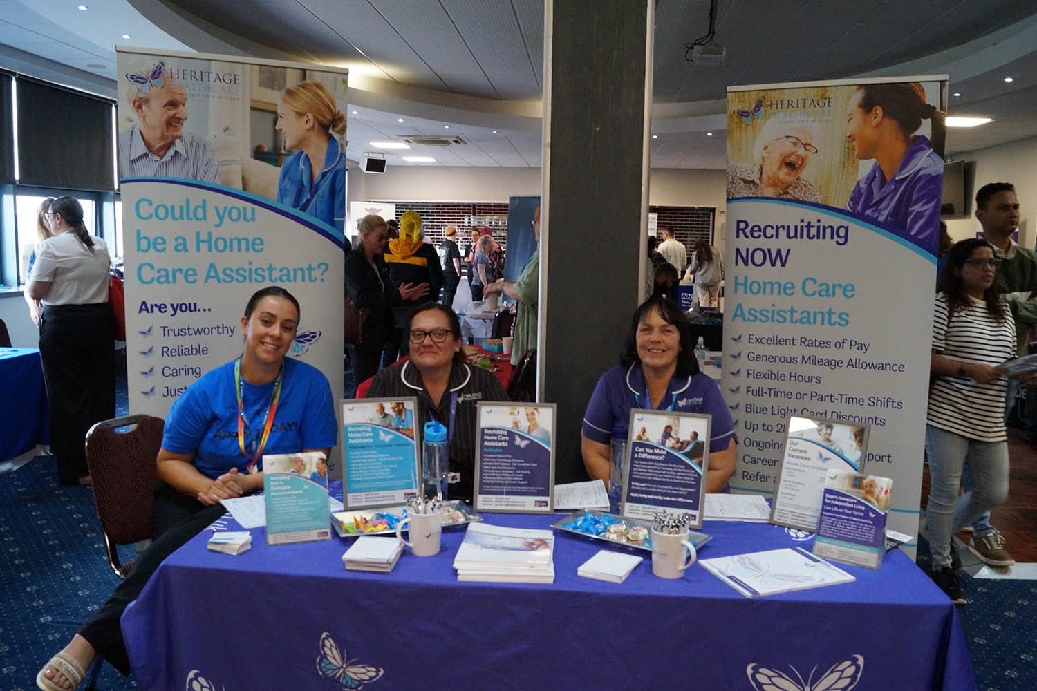 Registered Care Manager, Lisa, a white woman with dark hair tied up; she is wearing a black and white striped carer uniform and glasses. To the left of her is Kate from Make Care Matter, she is a white woman with brown tied up hair; she is wearing a blue t-shirt and dark trousers. On the right side of the table is Care Co-ordinator, Carol, a white woman with dark hair; she is wearing a purple carer tunic with white piping around the collar and the Heritage Healthcare logo on her left chest. They are both stood behind our stand at the careers fair. The stand includes a branded Heritage Healthcare tablecloth; leaflets showing our job opportunities, two branded cups with branded pens in, A6 and A4 branded notepads; sweets; and A4 posters (framed) showing our job opportunities. Behind the stand there are two banners promoting joining our team as a Home Care Assistant.