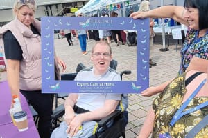 Client, Simon, a white man with short brown hair wearing a white t-shirt and grey bottoms, taking a selfie with Heritage Healthcare's selfie frame