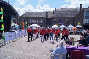 The 'Texas Toes Tappers,' four lines of older adults all female with one older man, performing at the GOLD Tea Dance. They are all wearing a red t-shirt with their groups name on the back.
