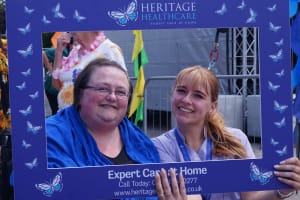 Client Kerami and her carer taking a photo with a Heritage Healthcare branded selfie frame. Kerami is a white woman with short dark hair wearing a black top with a blue jumper. 