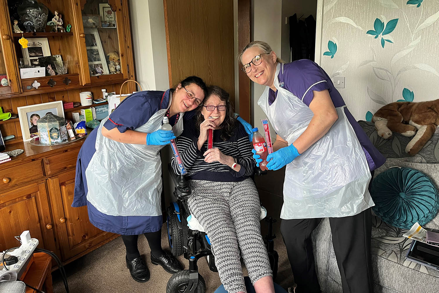 Two carers stood either side of a client in their home. They are all holding a cold drink as well as an ice pop. The carers are wearing purple tops with black bottoms and black shoes. On top of their shirts they have a medical apron on.