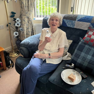 Client, a older white women with short curly hair, wearing a cream coloured shirt and blue trousers, sat on her couch with a cold drink.