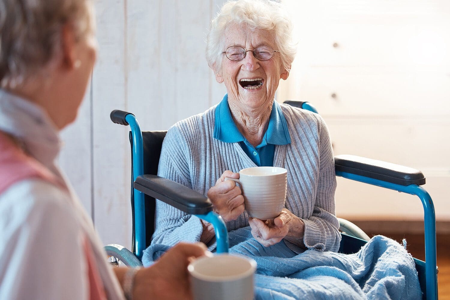 Elderly lady in a wheelchair enjoys a cup of tea with her carer. She is wrapped up with a blanket around her legs and is laughing