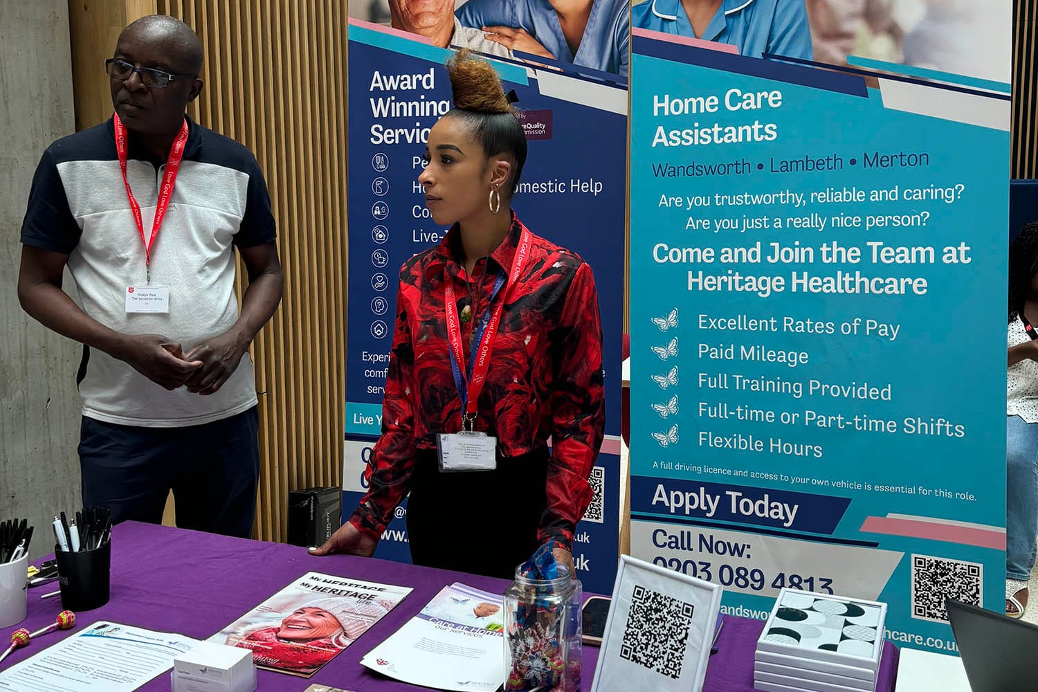 Michann and Sackson stood being the Heritage Healthcare stand at the event. Their is a table with a purple tablecloth on. On top of the the tablecloth there are pads, pens, business cards, a QR code to their website and some leaflets about their services. Behind the table there are two banners, one with recruitment information on and another with their services on