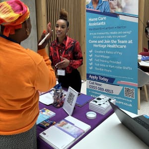 Registered Care Manager, Michann, a black woman with tired up dark hair, she is wearing a red top and black bottoms. Michann is talking to a jobseeker at the Heritage Healthcare stand. On the stand there is a QR code for their website, pens, notepads and a recruitment banner.