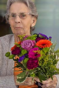 One of Heritage Healthcare Windsor' clients who won the draw. She is an old women with short grey hair wearing a knitted sweater, she is holding an arrangement of flowers that she won.