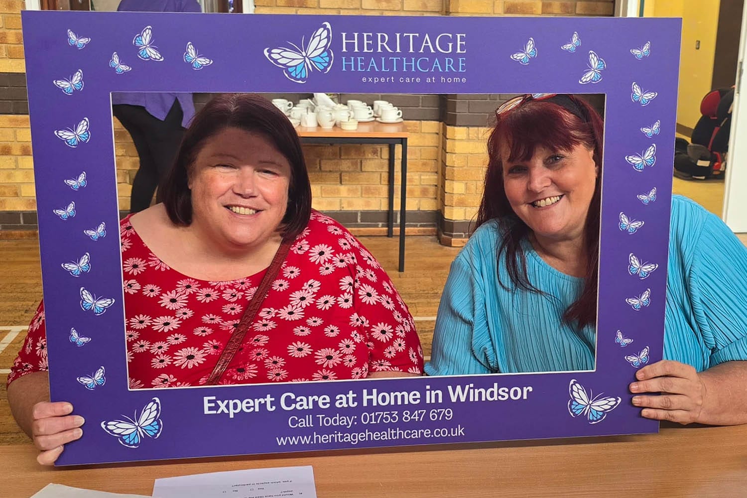 Two white women with red hair smiling whilst having their picture taken behind the Heritage Healthcare selfie frame. The woman on the left is wearing a red top with pink flowers on and the woman on the right is wearing a blue top.