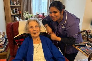 Carer, Peggy, a black women with medium length dark hair, wearing a purple Heritage Healthcare top and black bottoms. She is stood besides client, Peggy, who is an older white women with short grey hair, she is wearing a blue sweater and white shirt. Client Peggy is sat in a chair. They are both smiling at the camera.