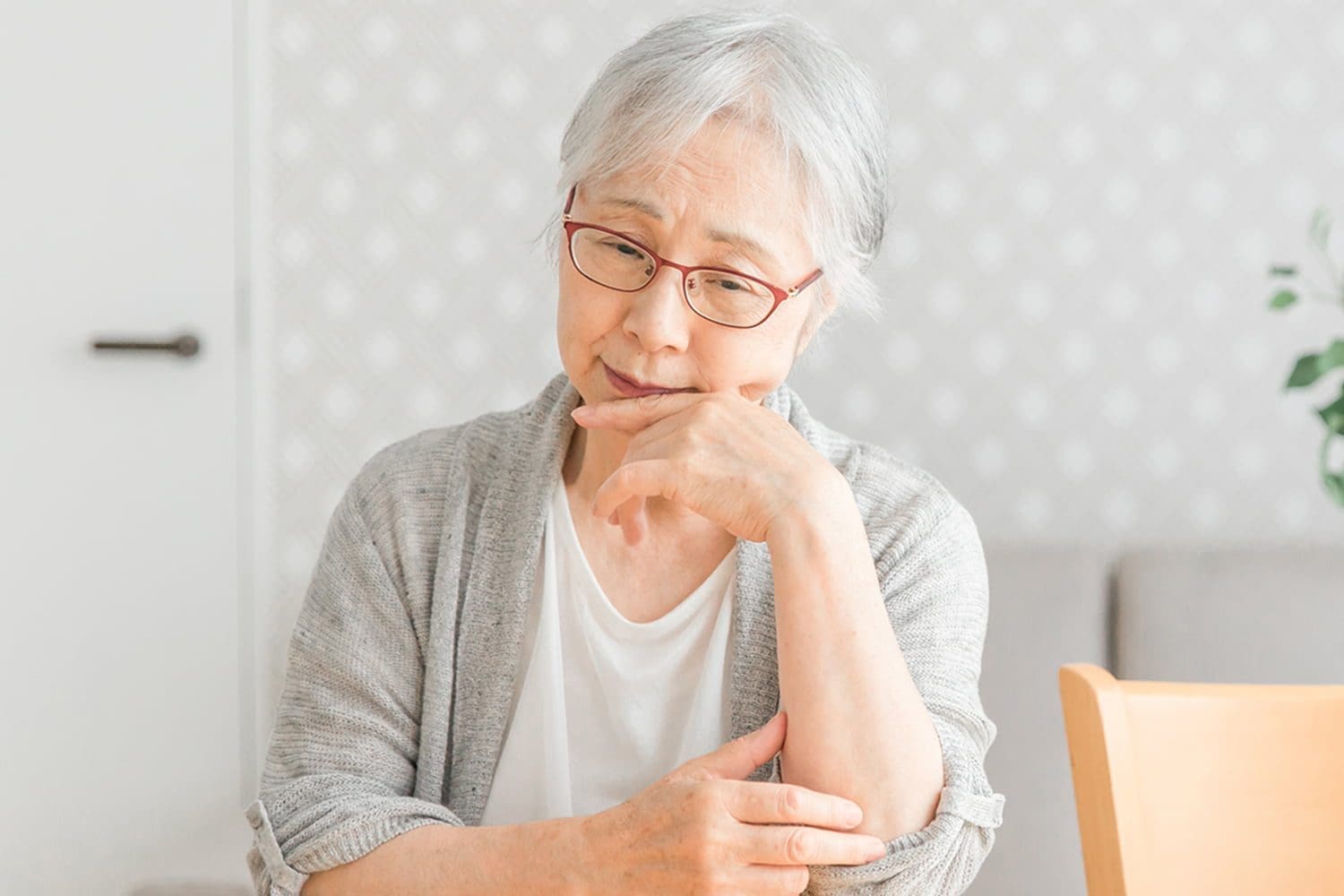 An elderly Asian lady with short grey hair wearing red glasses resting her head on her hand sitting at a table wearing a white t-shirt and grey cardigan.