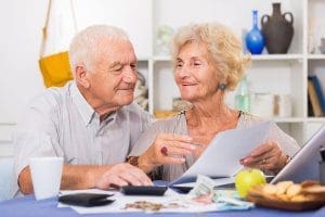 Elderly couple sat at a table with a laptop computer, money and documents in their home. Smiling at each other while pointing out information on a piece of paper