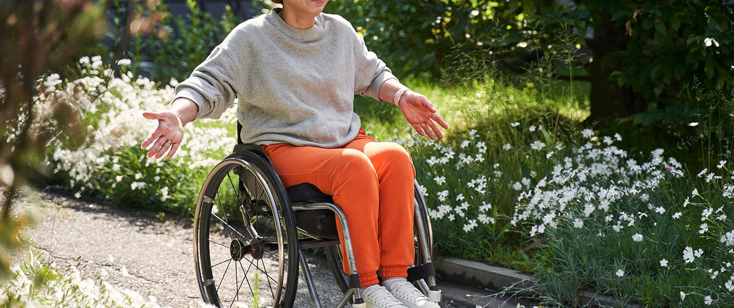 dark haired female wearing sunglasses, a grey jumper and orange trousers in a wheelchair going down a path surrounded by white flowers