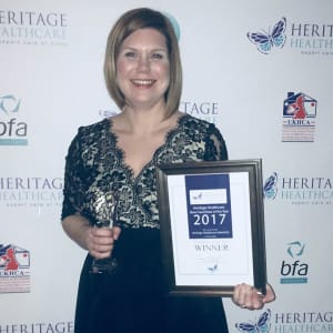 Marie Binks standing in front of a white wall, with Heritage Healthcare. bfa, and UKHCA logo on it, holding an award certificate and trophy.