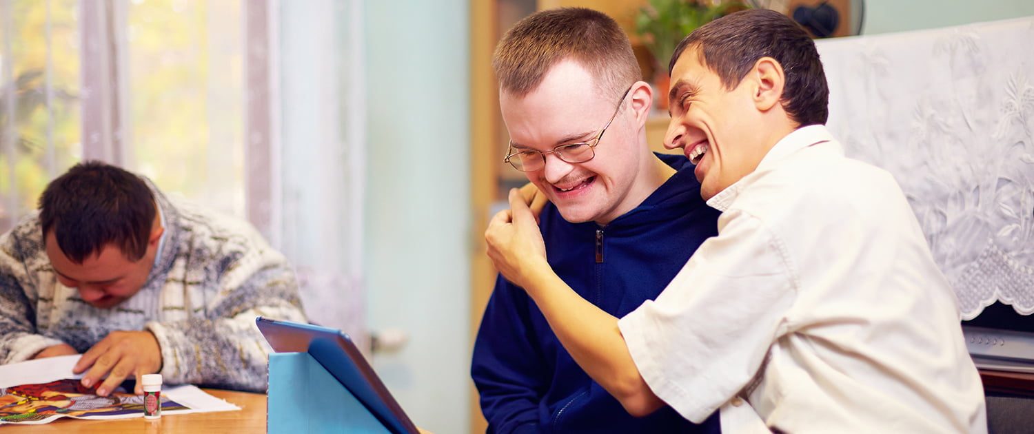 Male with downs syndrome being hugged by another disabled male while both looking at a computer tablet at a table