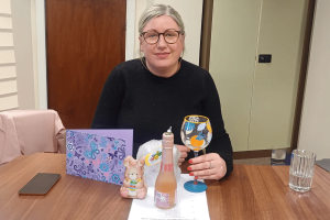 Carer, Marie, a white woman with long blonde hair, wearing a black sweater. She is sitting at a table with a thank you card, a chocolate bear and a bottle of prosecco in front of her.