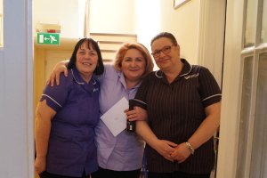 On the left is Care Co-ordinator, Carol. She is a white woman with short black hair wearing a purple Heritage Healthcare top and glasses. To the right of her is carer, Monika, a white woman with short blonde hair wearing a lilac Heritage Healthcare top and black trousers. Monika is also holding a card and bottle of prosecco To the right of her is Assistant Manager, Kay. A white woman with long brown hair. She is wearing a striped Heritage Healthcare top and glasses. All three are smiling at the camera.