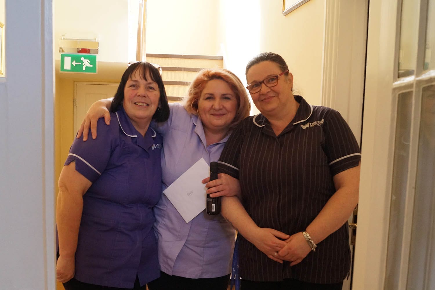 Carol, Monika and Kay smiling at the camera On the left is Care Co-ordinator, Carol. She is a white woman with short black hair wearing a purple Heritage Healthcare top and glasses. To the right of her is carer, Monika, a white woman with short blonde hair wearing a lilac Heritage Healthcare top and black trousers. Monika is also holding a card and bottle of prosecco To the right of her is Assistant Manager, Kay. A white woman with long brown hair. She is wearing a striped Heritage Healthcare top and glasses. All three are smiling at the camera.