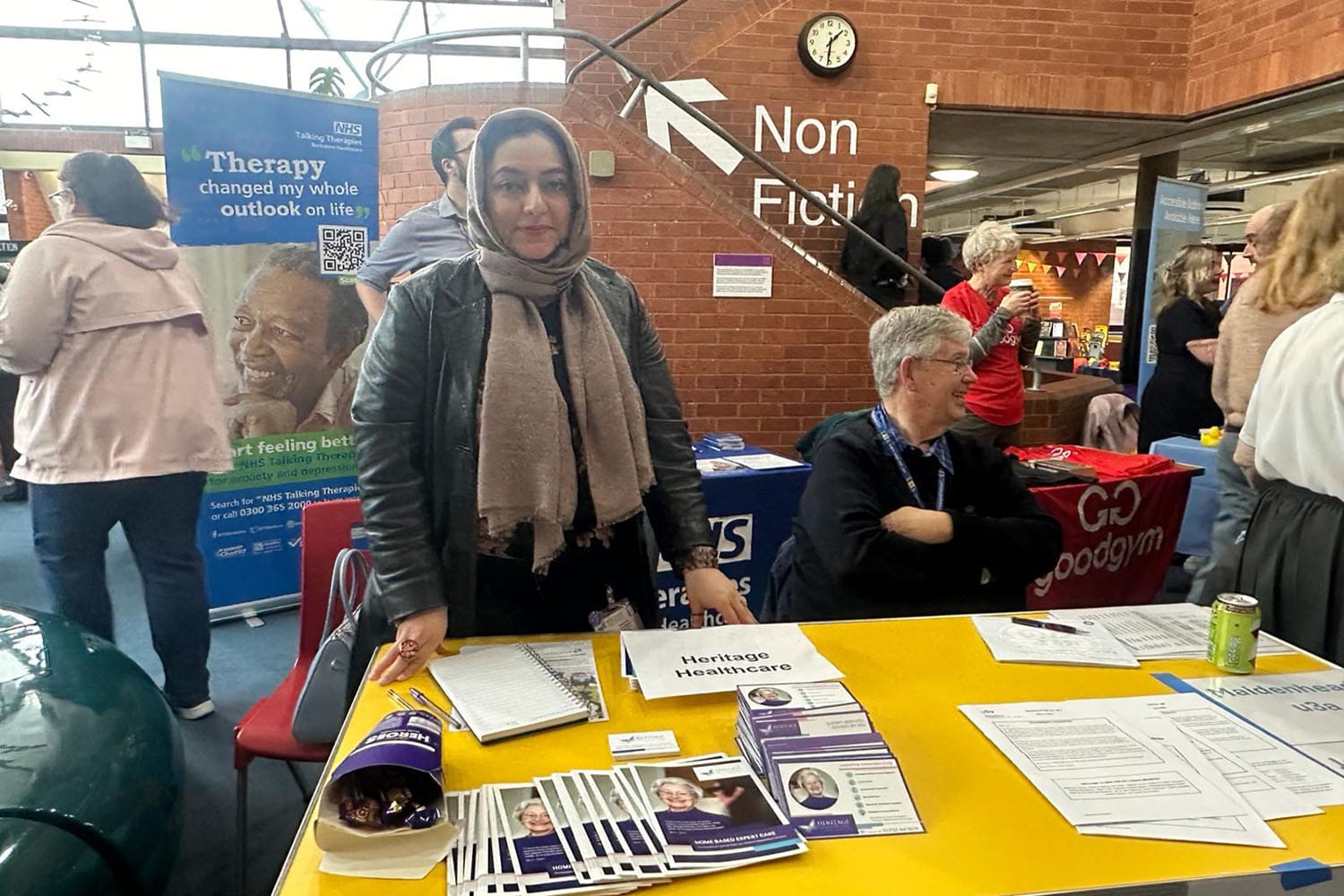 Care Manager, Sadaf, a brown women in a leather jacket, black floral dress and hijab. Stood behind Heritage Healthcare's stand at the RBWM (Royal Borough Windsor & Maidenhead) event