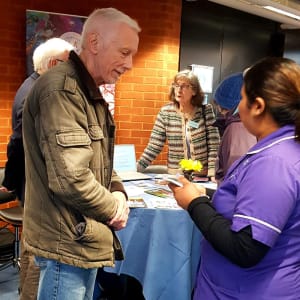Priya, a black women wearing a purple Heritage Healthcare top and black bottoms, talking to a white man with short white hair wearing a light brown coat and blue jeans.