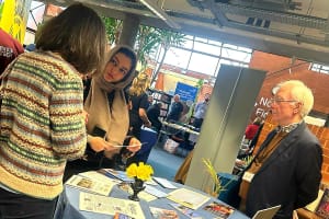 Care Manager, Sadaf, a brown women in a leather jacket, black floral dress and hijab, talking to a old white women whit short dark hair wearing a patterned sweater. With An old white man with short white hair in a brown jumper and blue shirt is stood to the side.