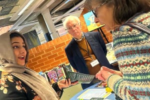 Care Manager, Sadaf, a brown women in a leather jacket, black floral dress and hijab, talking to a old white women whit short dark hair wearing a patterned sweater. An old white man with short white hair in a brown jumper and blue shirt is stood in the background