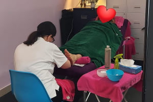 Heritage Healthcare carer covered with a green towel getting treatment on her feet. A white women with long black hair, wearing a white shirt and black trousers, providing treatment to a carers feet,