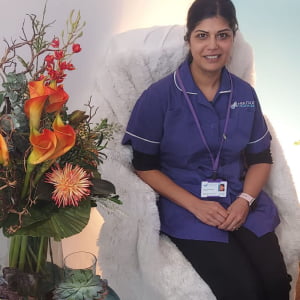 A brown woman with long dark hair, wearing a purple Heritage Healthcare top and black trousers, sat on a fluffy white chair. Next to her is a table covered in different types of plants