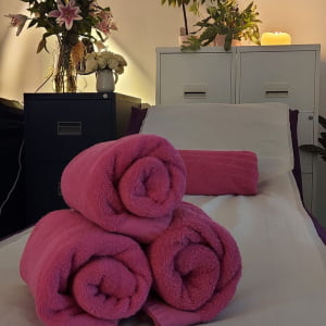 Three pink towels piled in a triangle shape, on top of a white massage bed. A fourth pink towel is laid towards the top of the bed. There are several plants and candles placed on cabinets in the background.
