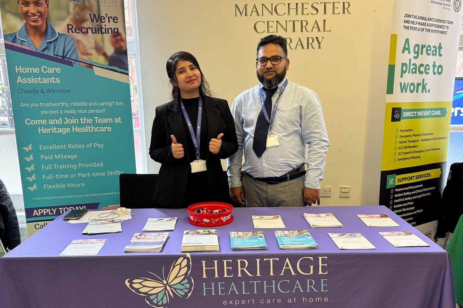 A dark skinned man and lady behind a table of leaflets with a purple tablecloth with the Heritage Healthcare logo. Lady is showing a thumbs up gesture