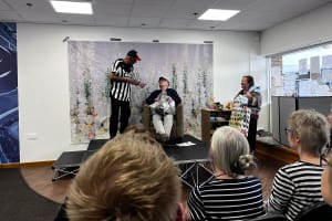 Performance of the short play 'Wor Grannie Joyce.' On the right side of the stage there is a black man wearing a Newcastle Football Club t-shirt with a black cap and black trousers. To the right of him is an older white man sit in a chair wearing grey trousers, a black jumper and a checked flat cap.