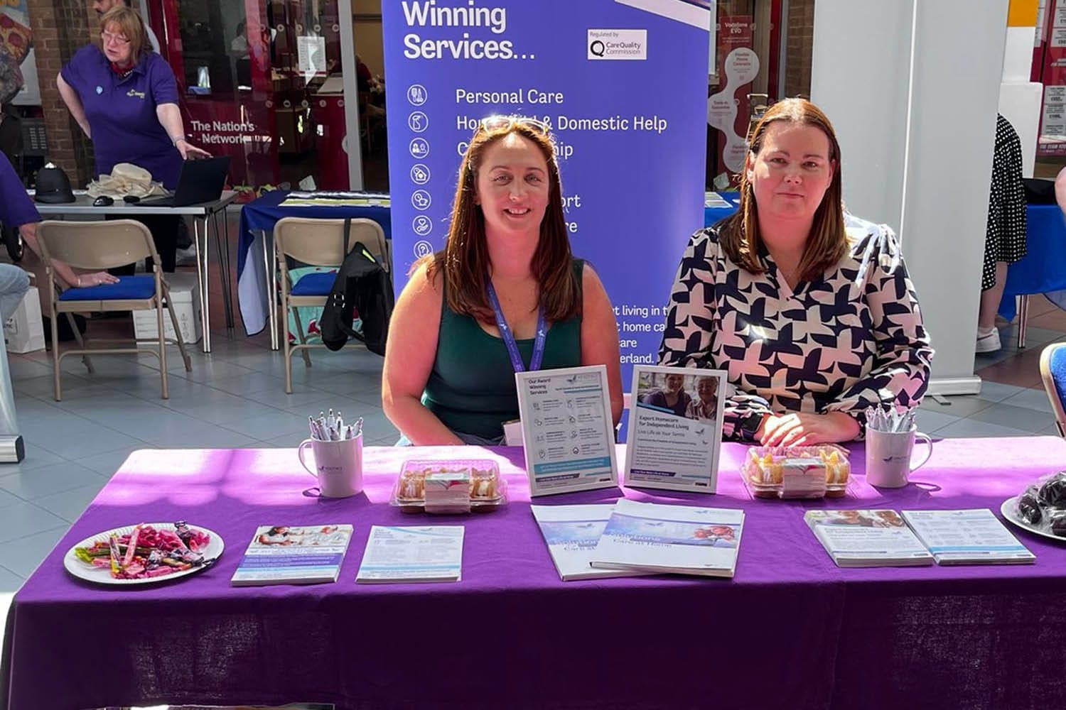 Heritage Healthcare's stall at the Dementia Roadshow Directors and Registered Care Managers, Suzanne and Tanya, sat behind their stall at the Dementia Roadshow. Their stall has a purple table cloth on with sweets, pens, cups and informational literature about their services.