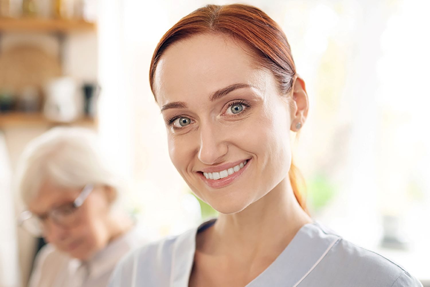 White female care worker with ginger hair and green eyes with an elderly lady in the background. She wears grey scrubs and a big smile