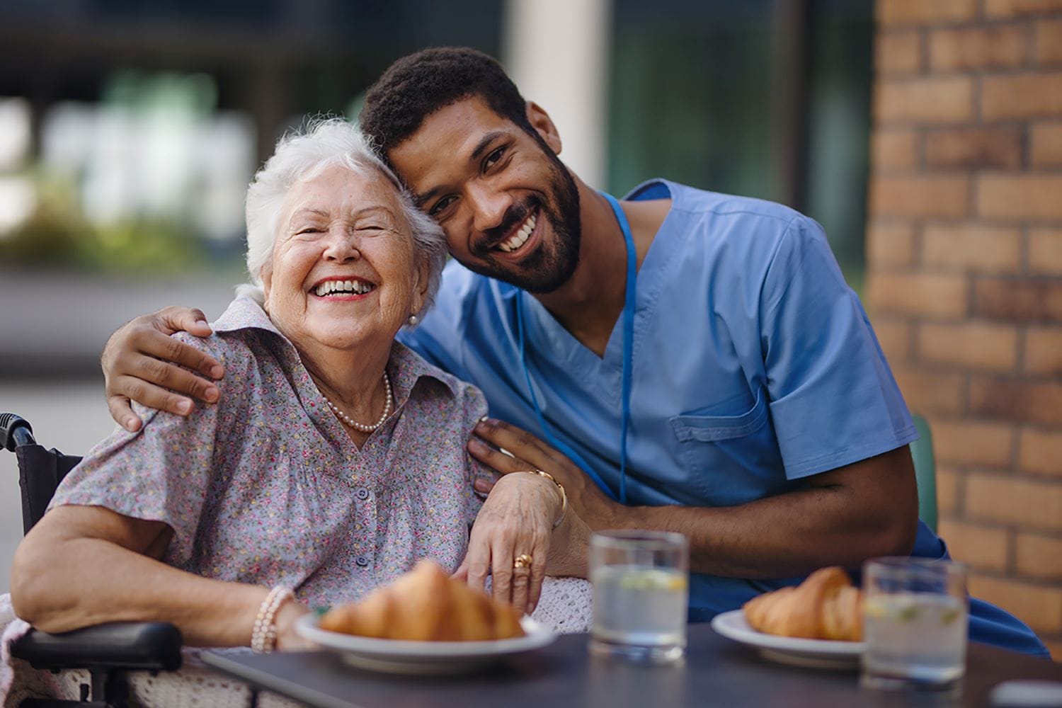Black male carer wearing blue scrubs hugging and elderly white lady with grey hair wearing a floral blouse. They are sitting at a table with drinks and croissants