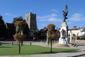 Photo of a memorial in Colchester. It has black statues on the bottom and top of a pillar. In the background there is a church,