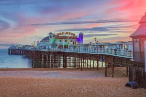 Photo taken from a beach of the Brighton Palace Pier entrance, during a sunset. The piers name is written in yellow curvy writing. Behind the entrance you can see festival/carnival rides.
