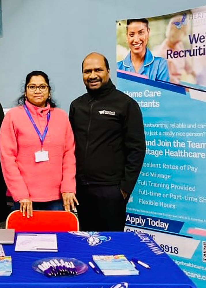 Directors, Deepti and Umakanth, as well as Registered Care Manager, Hannah, standing behind a recruitment and services display at Wellington Street Job Centre, Leicester. The display highlights award-winning services, including personal care and respite support. The table is set with informational brochures, a laptop, and snacks.