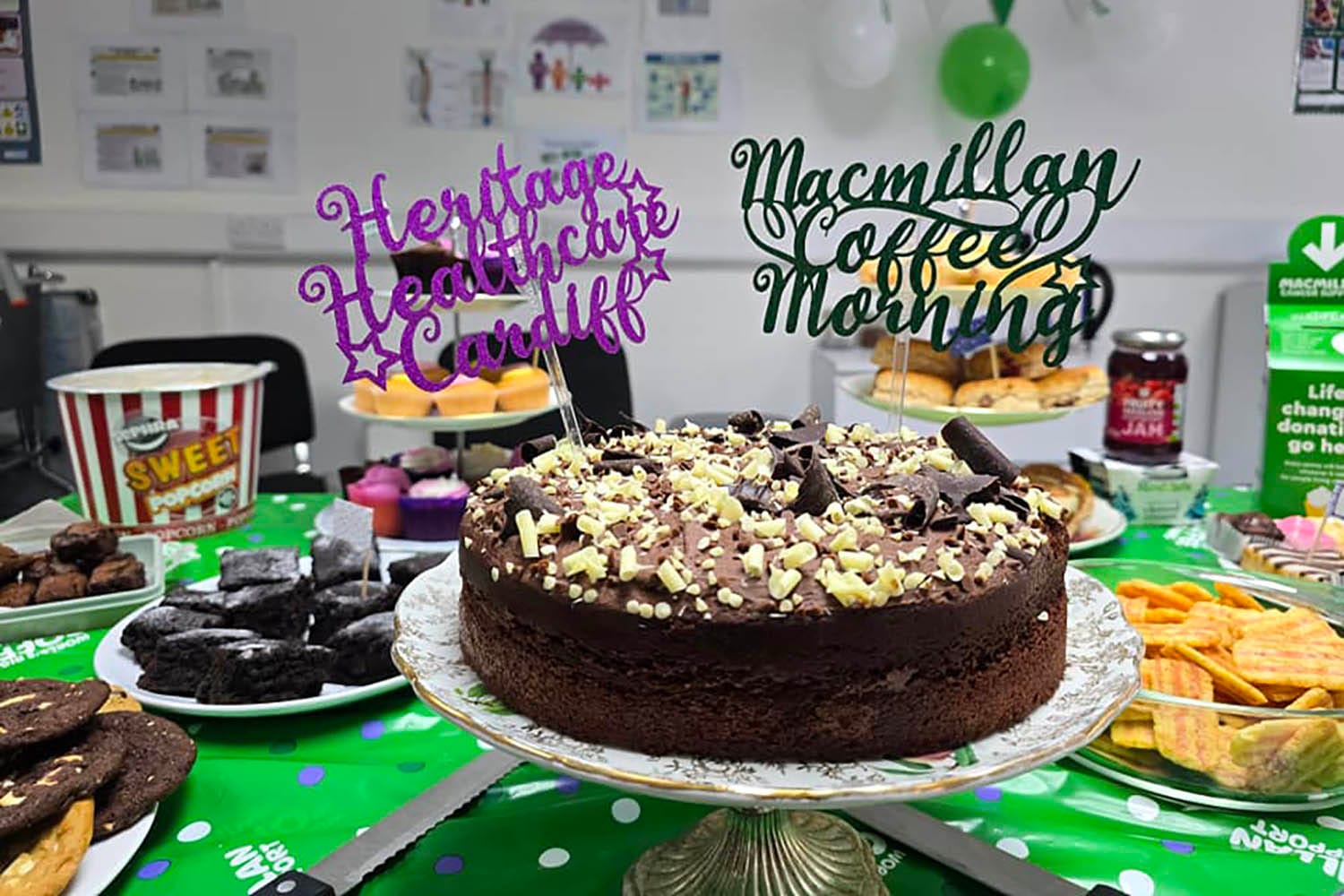 Close-up of a table filled with assorted baked goods and a centrepiece chocolate cake, decorated with white and dark chocolate shavings. Signs reading 