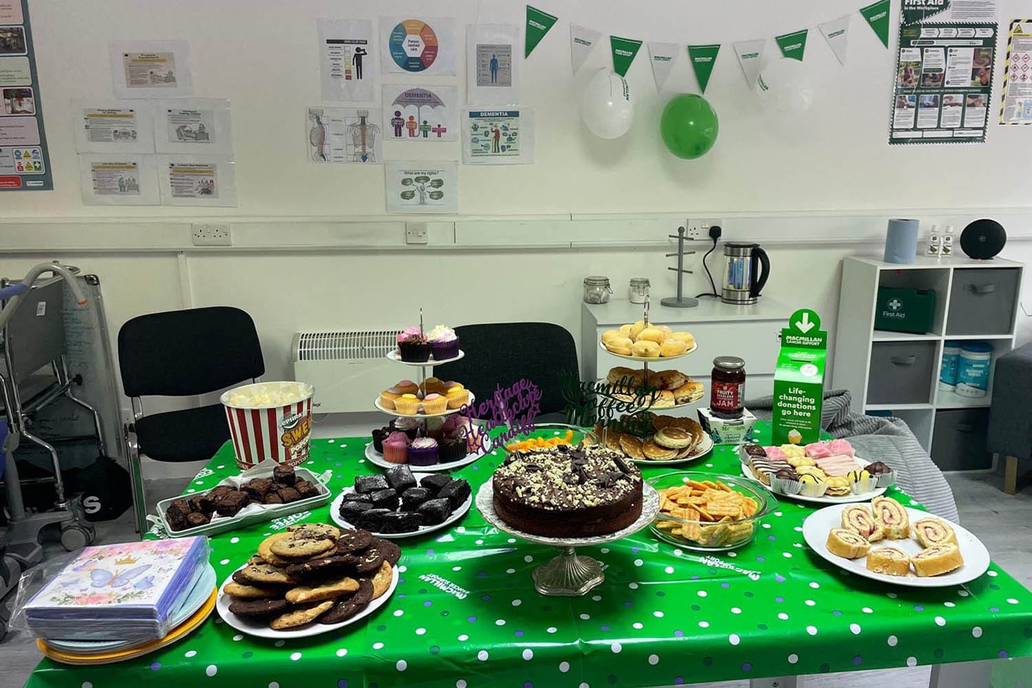 A variety of baked goods are laid out on a green MacMillan Coffee Morning tablecloth, including a tiered display of cupcakes, chocolate cake, popcorn, and jam. In the background, decorations such as balloons and flags in green and white highlight the event, while a donation box stands prominently on the table.