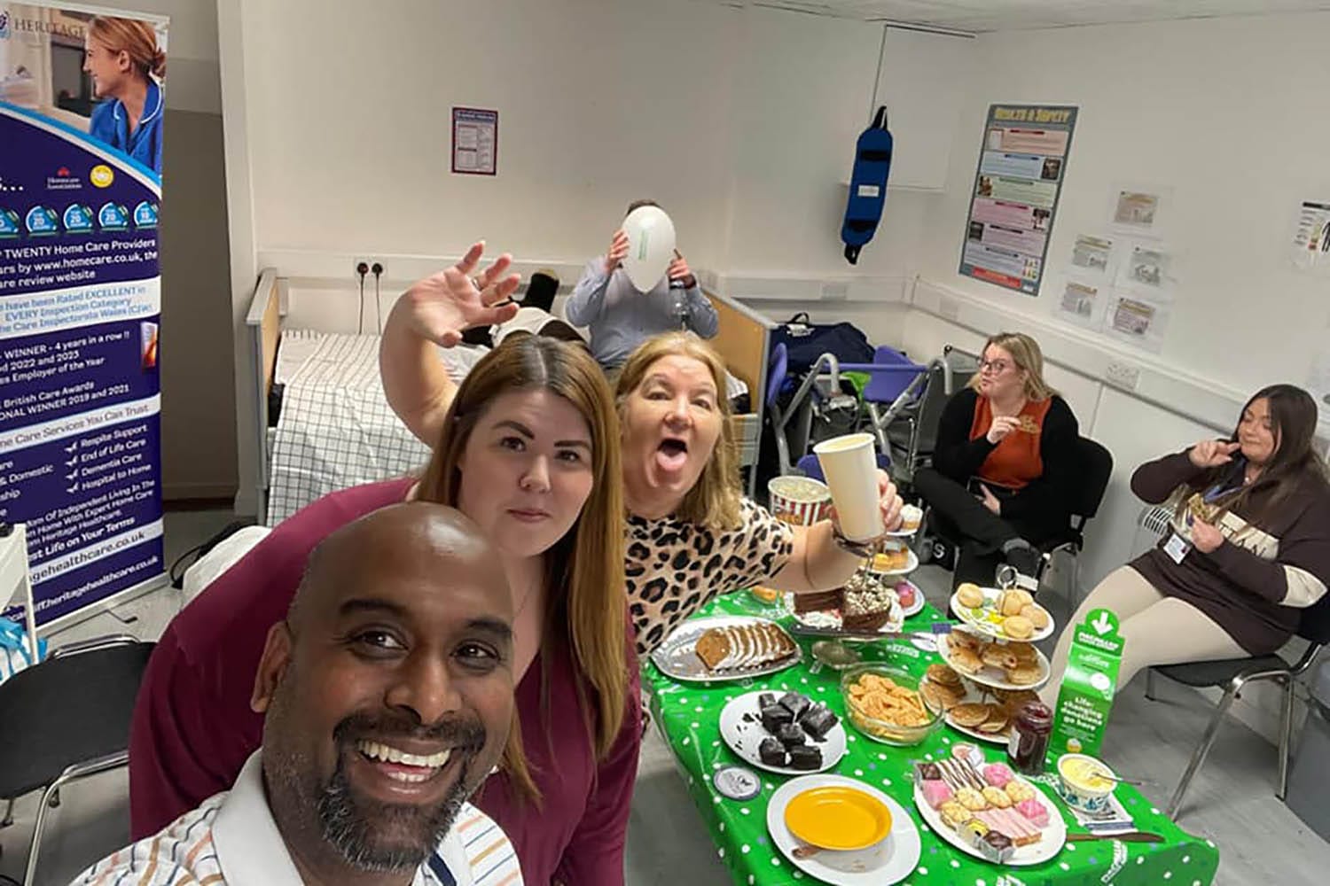 A group of people in a room enjoying a variety of snacks on a table covered with a green polka dot tablecloth. One person is taking a selfie, while others smile and hold drinks. Posters, promoting MacMillan and Heritage Healthcare Cardiff, as well as chairs are visible in the background.