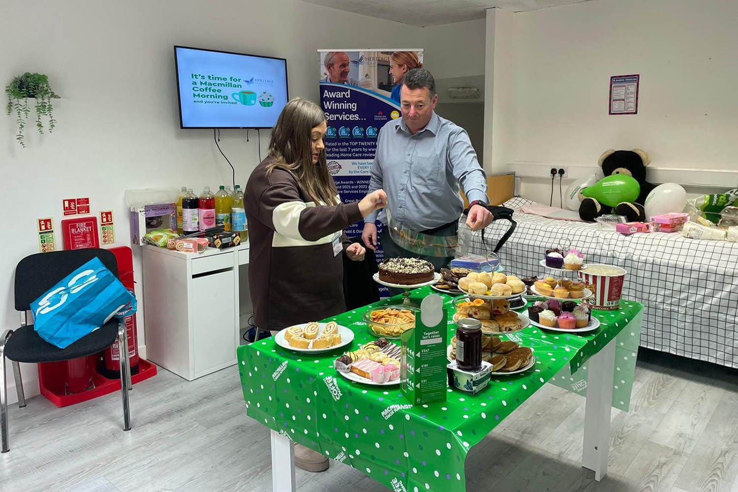 Two people are selecting food at a table set for a MacMillan coffee morning event. A woman wearing a brown and white sweater and a man in a blue shirt are seen reaching for pastries and desserts. The table is covered with a green tablecloth featuring the MacMillan logo, and it holds a variety of baked goods such as cakes, muffins, and rolls. A TV in the background displays a sign for the MacMillan Coffee Morning, and informational posters are visible on the walls. In the corner, a plush bear is resting on a bed with a green MacMillan balloon.