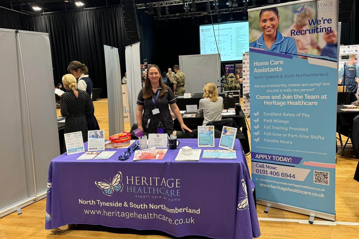 Director, Suzanna, a female with long light brown hair and pale blue eyes wearing a Heritage Healthcare top, stood behind the Heritage Healthcare North Tyneside stand at the Northumbria University Part-time Job Fair. The stand had a branded tablecloth with sweats, leaflets, lanyards, and framed photos advertising the jobs they had on offer. Behind the stand, there is a banner promoting their jobs as well.