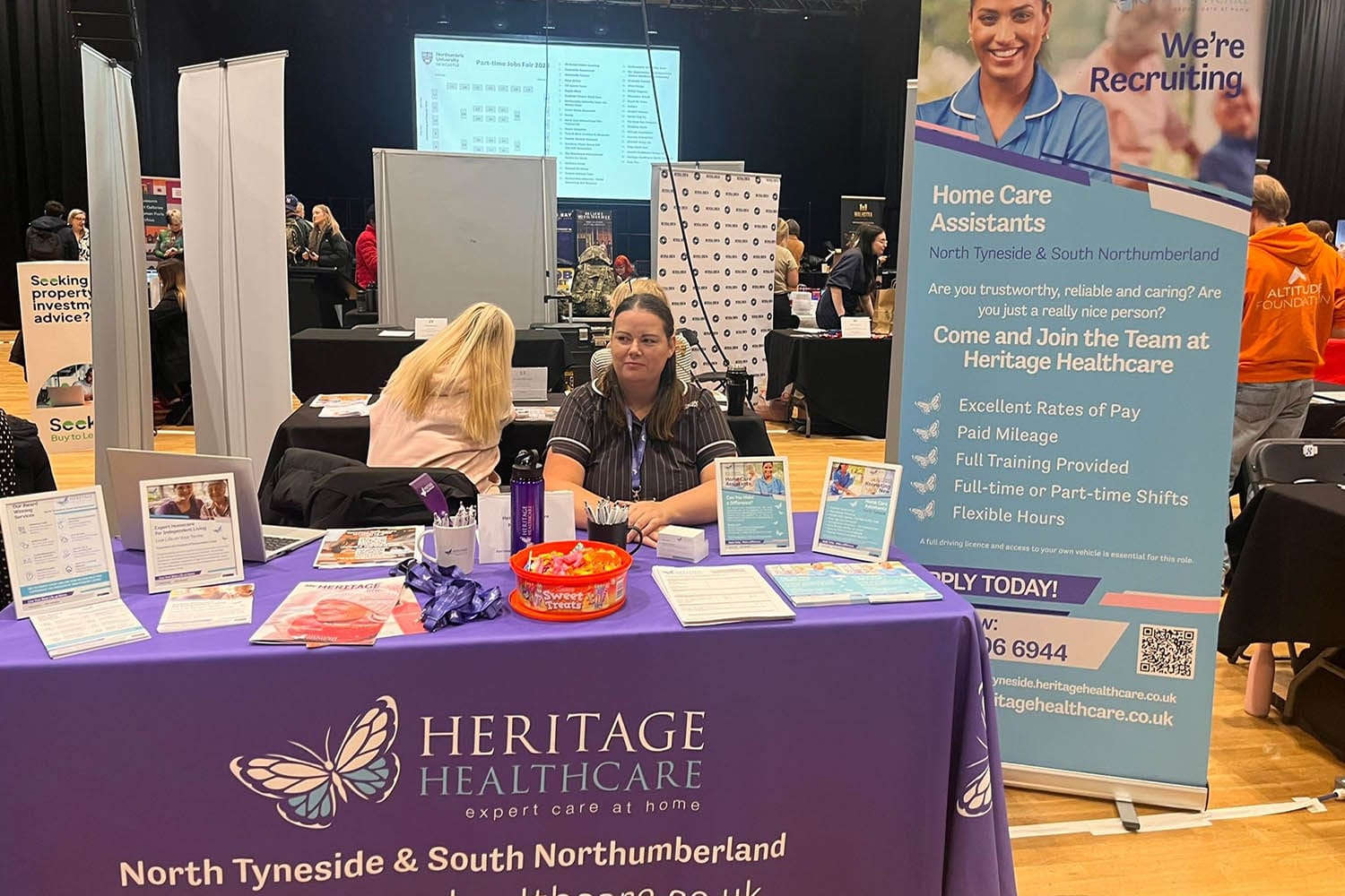 Director, Tanya, a female with long dark hair and brown eyes wearing a Heritage Healthcare top, sat behind the Heritage Healthcare North Tyneside stand at the Northumbria University Part-time Job Fair. The stand had a branded tablecloth with sweats, leaflets, lanyards, and framed photos advertising the jobs they had on offer. Behind the stand, there is a banner promoting their jobs as well.