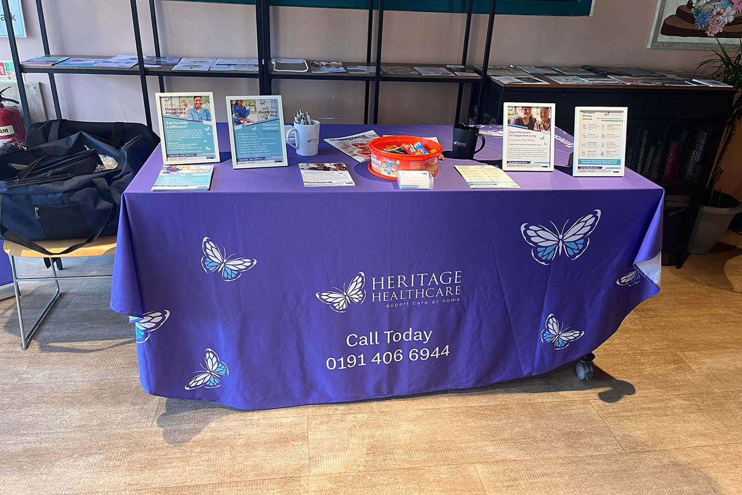 Table covered with a purple, branded Heritage Healthcare tablecloth. On the table are brochures and informational flyers about the services Heritage Healthcare North Tyneside offers; a mug; and a red bowl filled with chocolates.