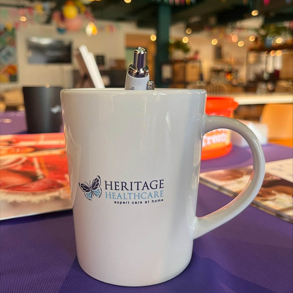 A white Heritage Healthcare branded coffee mug with a Heritage Healthcare branded pen in it. The mug is on a table surrounded by brochures and informational flyers.