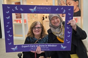 Two smiling women pose inside a large, purple selfie frame with "Heritage Healthcare" and butterflies on it. They are in Nicholsons Shopping Centre. There are various products displayed in the background.