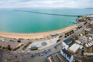 Aerial view of a Southend-on-Sea with a sandy beach and a view of Southend pier extending into the sea. The promenade has a circular structure and several parked cars. Buildings line the shoreline, and the sky is overcast.